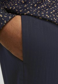 Pantalons bleu marine à rayures avec une texture lisse, partiellement recouverts par une chemise sombre à motifs avec de petits motifs floraux dorés.