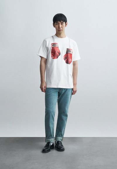 Λευκό κοντομάνικο t-shirt με γραφικό σχέδιο κόκκινων γαντιών πυγμαχίας. Συνδυασμένο με ανοιχτόχρωμο τζιν τζιν και μαύρα λουστρίνια παπούτσια. Λείο ύφασμα.