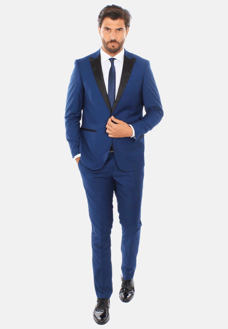 Tuxedo blu con revers in satin nero, design slim-fit, abbinato a una camicia bianca e una cravatta blu navy. Scarpe eleganti nere lucide.
