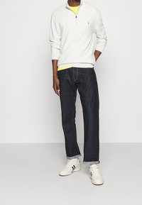 Weißer Zip-Kragen-Pullover, über einem gelben Hemd getragen, kombiniert mit dunkelblauen Jeans und weißen Turnschuhen mit grünen Akzenten.