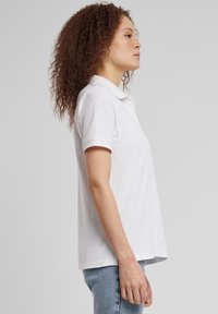 Bijela polo majica s mekanom teksturom, kratkim rukavima i klasičnim ovratnikom. Bočni pogled prikazuje opušteni kroj i glatki rub.