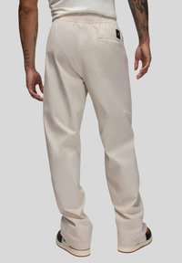 Beigare bomullsbyxor med elastisk midjeresår, raka ben och bakficka. Har sömnadsdetaljer och en logotypmärke på midjeresåren.