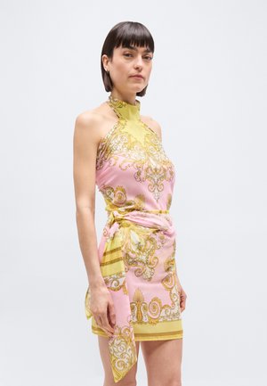 Rövid sötét hajú nő ujjatlan, diplomata nyakú, rózsaszín és sárga barokk mintás ruhában, kötött derékszalaggal.