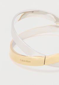 Silber- und goldfarbener, gedrehter Armreif mit polierter Oberfläche. Enthält das gravierte "Calvin Klein"-Logo im goldenen Teil.