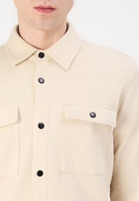 Bej színű, gombos ing, texturált anyagból készült, gallérral, két elülső zsebbel fekete gombokkal, és letisztult, minimalista dizájnnal.