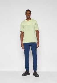 Ανοιχτό πράσινο αθλητικό μπλουζάκι με υφασμένο σχέδιο σε συνδυασμό με μπλε σκούρο παντελόνι cargo με τσέπες και λογότυπο, φορεμένο με μαύρα αθλητικά παπούτσια.