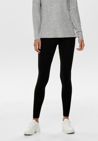 Leggings noirs en matériau extensible, coupe ajustée, assortis à un haut à manches longues gris chiné et des chaussures de sport blanches.