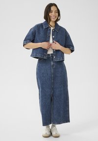 Lyhythihainen cropattu farkkutakki nappikiinnityksellä, yhdistettynä pitkään farkkuhameeseen. Molemmat vaatteet tummansiniset ja haalistuneella tekstuurilla.