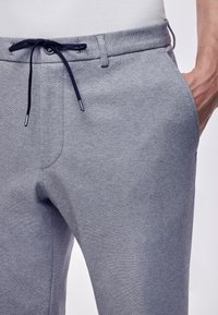 Šedé tepláky s hladkou strukturou, se širokým elastickým pasem a nápletem v námořnické modři. Obsahují boční kapsy a mají uvolněný střih.