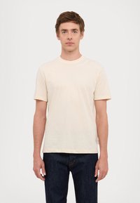 T-shirt di cotone beige chiaro con maniche corte, scollatura rotonda e dettaglio del logo bianco sulla spalla sinistra. Abbinato a jeans di denim blu scuro.