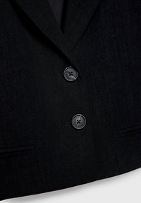 Blazer noir texturé avec deux grands boutons noirs et un col à revers cranté, montrant une partie des poches avant.