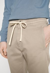 Beige katoenen broek met een elastische tailleband, trekkoordsluiting en zijzakken. Vergezeld van een lichtblauwe shirt met lange mouwen.