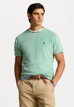 Lichtgroene badstof t-shirt met een ronde hals en korte mouwen, met een klein marineblauw logo. Gedragen over een gestreept hemd.