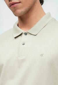 Világos bézs pólóing, amely texturált pamutból készült. Fitted gallérral, három gombbal és finoman hímzett logóval rendelkezik a mellkason.