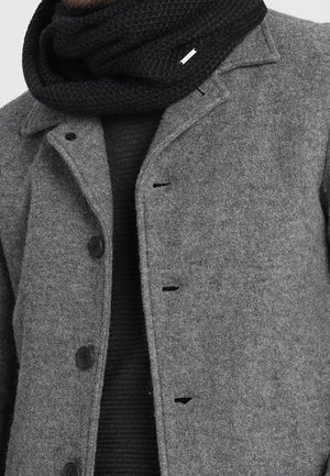 Szary wełniany płaszcz z czarnymi guzikami na czarnym prążkowanym swetrze, w połączeniu z grubym czarnym dzianym szalem owiniętym wokół szyi.