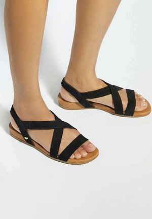 Voeten met zwarte platte sandalen met bandjes en bruine zolen op een witte achtergrond, die een zij- en voorkant van het schoeisel tonen.