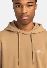 Tan hoodie made of soft fabric, featuring a kangaroo pocket, drawstrings, and white printed logo on the chest.