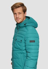Teal pufferjack met een capuchon, met een rits aan de voorkant, twee borstzakken en een logo patch op de linkermouw. Horizontale quilt-details.