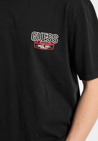 T-shirt nera in cotone con logo ricamato rosso e bianco "GUESS Originals" e "SINCE 1981" sotto, maniche corte.