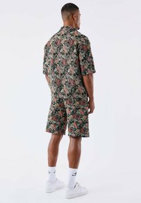 Chemise à manches courtes à imprimé floral et short assorti en vert foncé avec des roses roses et crème. Porté avec des baskets blanches et des chaussettes courtes.
