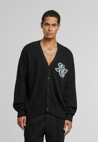Svart strikket cardigan med dyp V-hals, som har en enkelt frontlomme med et hvitt brodert design og fem knapper foran.