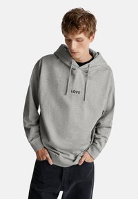 Giovane uomo in felpa grigia con cappuccio e la scritta "LOVE", mani nelle tasche, che guarda in basso, indossando pantaloni scuri su sfondo bianco.