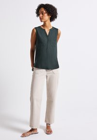 Ciemnozielony top bez rękawów z dekoltem w serek i dwiema kieszeniami na piersi, zestawiony z jasnobeżowymi spodniami oraz minimalistycznymi srebrnymi sandałami.
