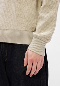 Pulover bej cu margini ondulate, având un finisaj texturat, care prezintă un manșon și o margine ajustate. Mâna se sprijină pe o pereche de pantaloni din denim închis.