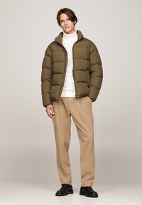 Doudoune vert olive avec fermeture éclair, col montant et texture matelassée, portée sur un pull blanc et un pantalon beige.