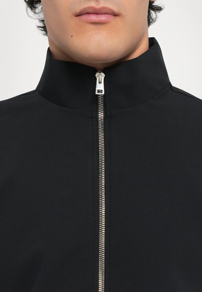 Svart zip-up jakke med høy krage, laget av glatt materiale. Har en sølvglidelås og et minimalistisk design.