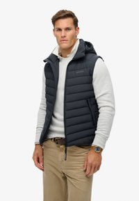 Marineblå puffervest med hætte og horisontalt quiltet design, båret over en lysegrå sweater og parret med tanfarvede bukser og et brunt bælte.