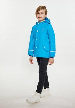 Helle blaue wasserdichte Jacke mit Kapuze, ausgestattet mit weißen reflektierenden Streifen, vorderen Knöpfen und einer seitlichen Tasche. Über dunklen Hosen und weißen Schuhen getragen.