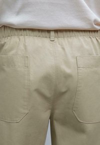 Pantalons en coton beige avec une taille élastique, équipés de deux poches arrière et d'une texture lisse. Design simple avec des détails de couture minimalistes.