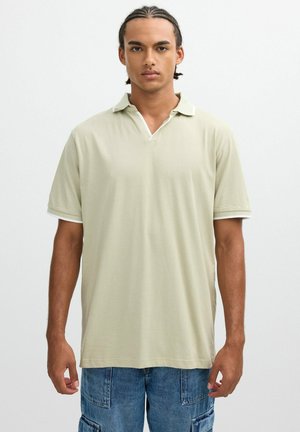 Joven con cabello trenzado que lleva puesta una camiseta polo beige de manga corta y pantalones cortos cargo vaqueros azules, de pie frente a un fondo blanco liso.