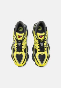 Black and yellow sneakers feature a mesh upper, reflective accents, and a thick black sole with red detailing. Unique design lines enhance visibility.