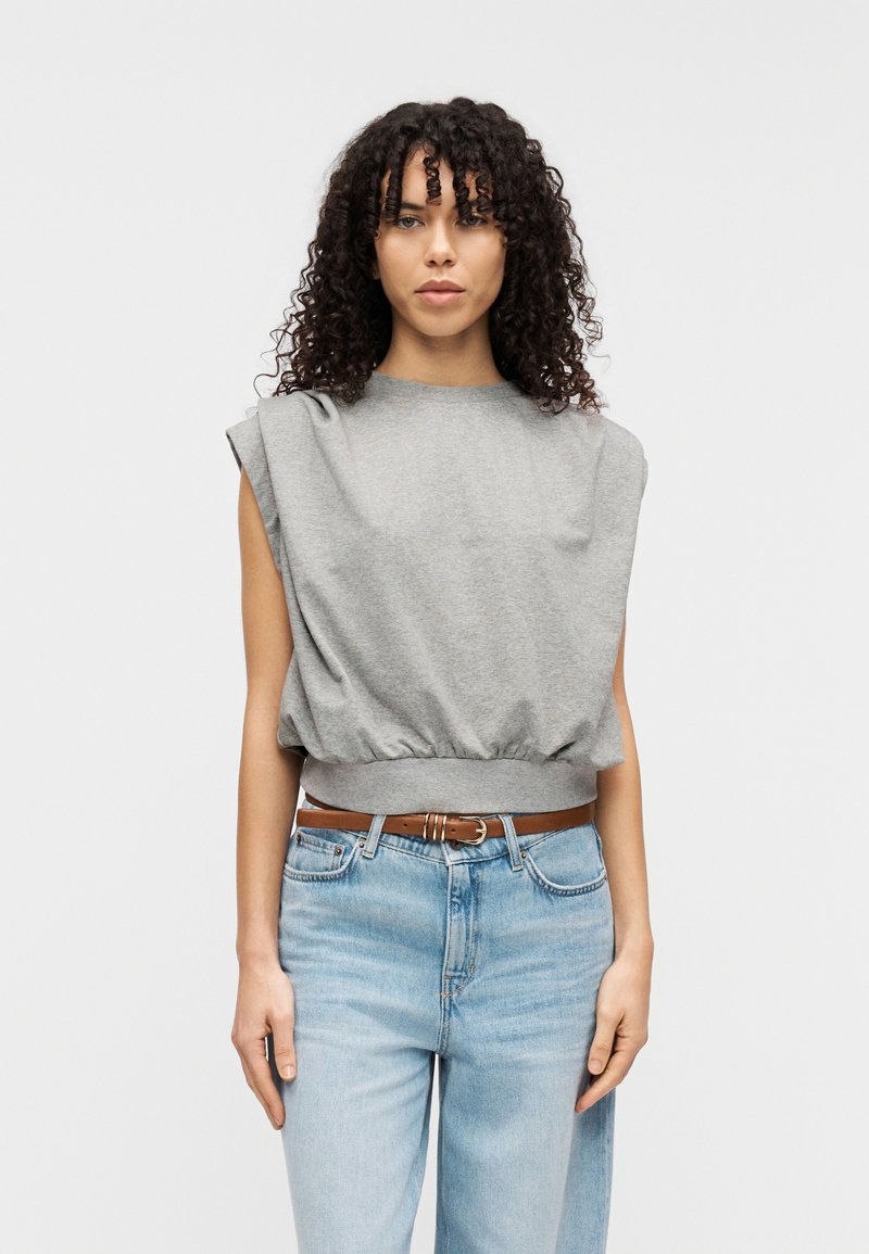 Grå ermeløs topp med avslappet passform, rynket kant og myk tekstur. Kombinert med lyseblå denimjeans og brunt belte.