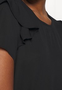 Schwarze Bluse aus Stoff mit kurzen Rüschensäumen, glatter Textur und rundem Ausschnitt, die ein einfaches, aber elegantes Design hervorhebt.