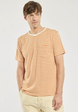 Homme portant un T-shirt à manches courtes rayé orange et blanc avec une poche poitrine, pantalon beige, debout devant un fond uni, mains dans les poches.