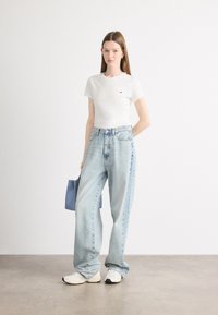 Camiseta de algodón blanca, pantalones vaqueros de mezclilla sueltos de color azul claro y zapatillas blancas. Sosteniendo un pequeño bolso azul, en un fondo claro.