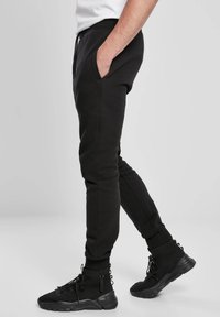 Fekete, puha anyagból készült melegítőnadrág szűkített szárakkal, rugalmas mandzsettákkal és oldalzsebekkel, fekete magas szárú tornacipőkkel párosítva, melyeken fűződíszítés található.