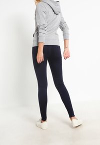 Felpa grigia con cappuccio, maniche lunghe e polsini a costine, abbinata a leggins blu scuri e sneaker bianche, vista da dietro.