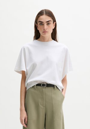 Jonge vrouw staat recht voor zich uit kijkend, draagt een eenvoudige witte losse korte mouwen shirt, ingestopt in een hoog getailleerde olijfgroene broek met een zwarte riem.