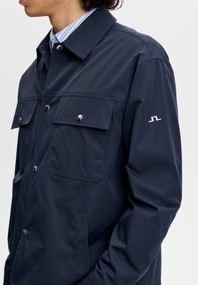 Uomo che indossa una giacca blu navy con bottoni, con tasche a patta sul petto e piccolo logo bianco sulla manica sinistra, sopra una camicia a righe con colletto.