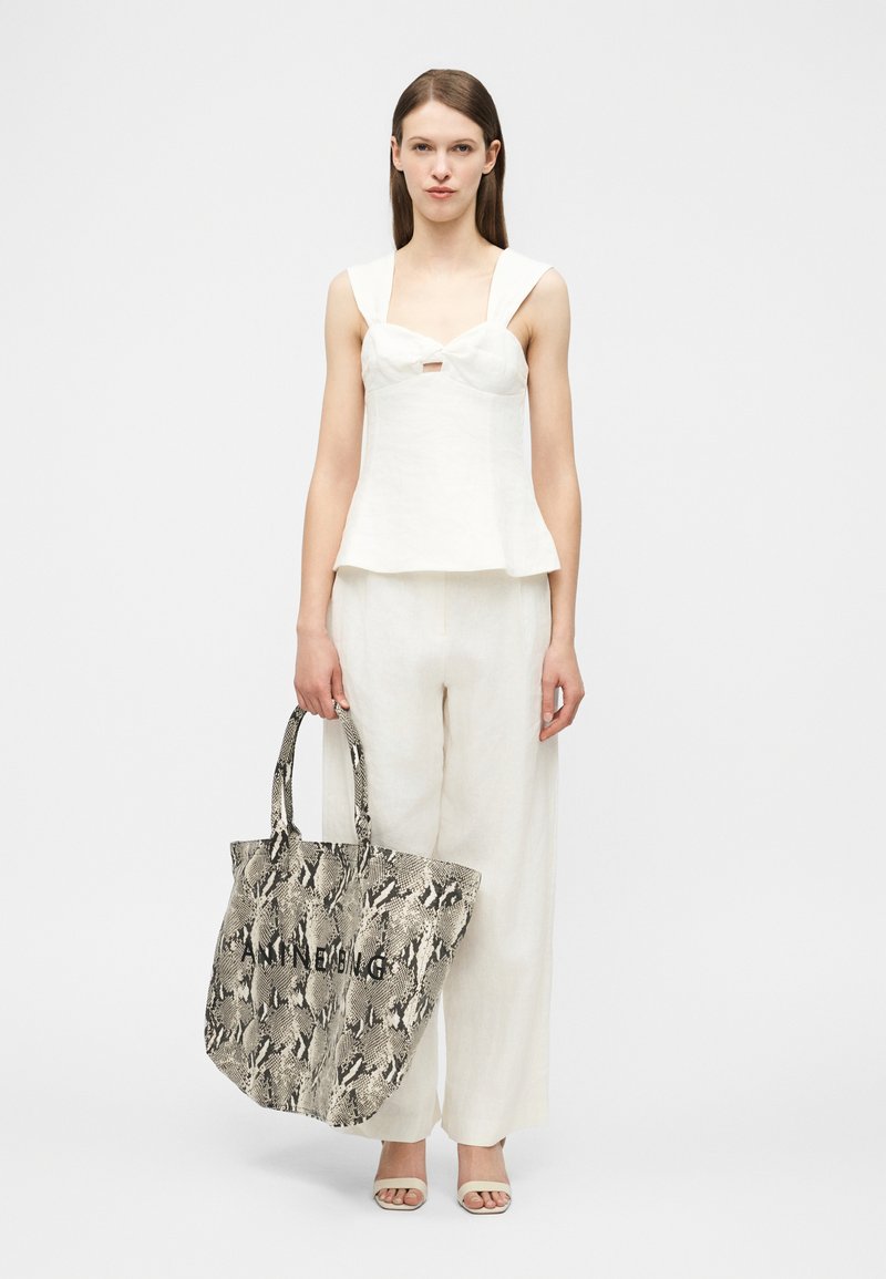 Γυναίκα που φοράει λευκή αμάνικη μπλούζα και παντελόνι, κρατώντας μια μεγάλη τσάντα ώμου με σχέδιο δέρματος φιδιού, στέκεται μπροστά σε μια απλή φόντο.