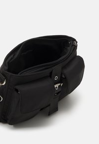 Borsa nera in nylon con chiusura a zip, dotata di una tasca esterna e di una tracolla con dettagli metallici. Interno foderato con materiale morbido.