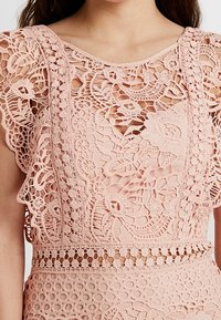 Světle růžový krajkový top s jemnými květinovými vzory, volánkovými rukávy a vykrojeným výstřihem s oblikem klíčové dírky.