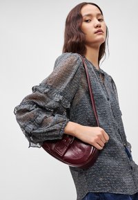 Κόκκινη δερμάτινη τσάντα χιαστί με καμπυλωτό σχήμα, δομημένο κλείσιμο και ιμάντα ώμου, συνδυασμένη με ένα μοτίβο, διάφανο μπλουζάκι με μακρύ μανίκι.