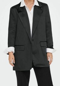 Veste blazer noire oversize en tissu lisse avec un seul bouton, des revers crantés et des poches plaquées à l'avant. Présente des poignets de chemise blancs contrastants.