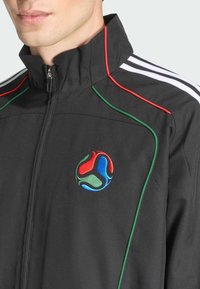 Sort zip-up jakke med røde og grønne accenter, med et cirkulært flerfarvet broderet logo på brystet og hvide stribede skuldre.