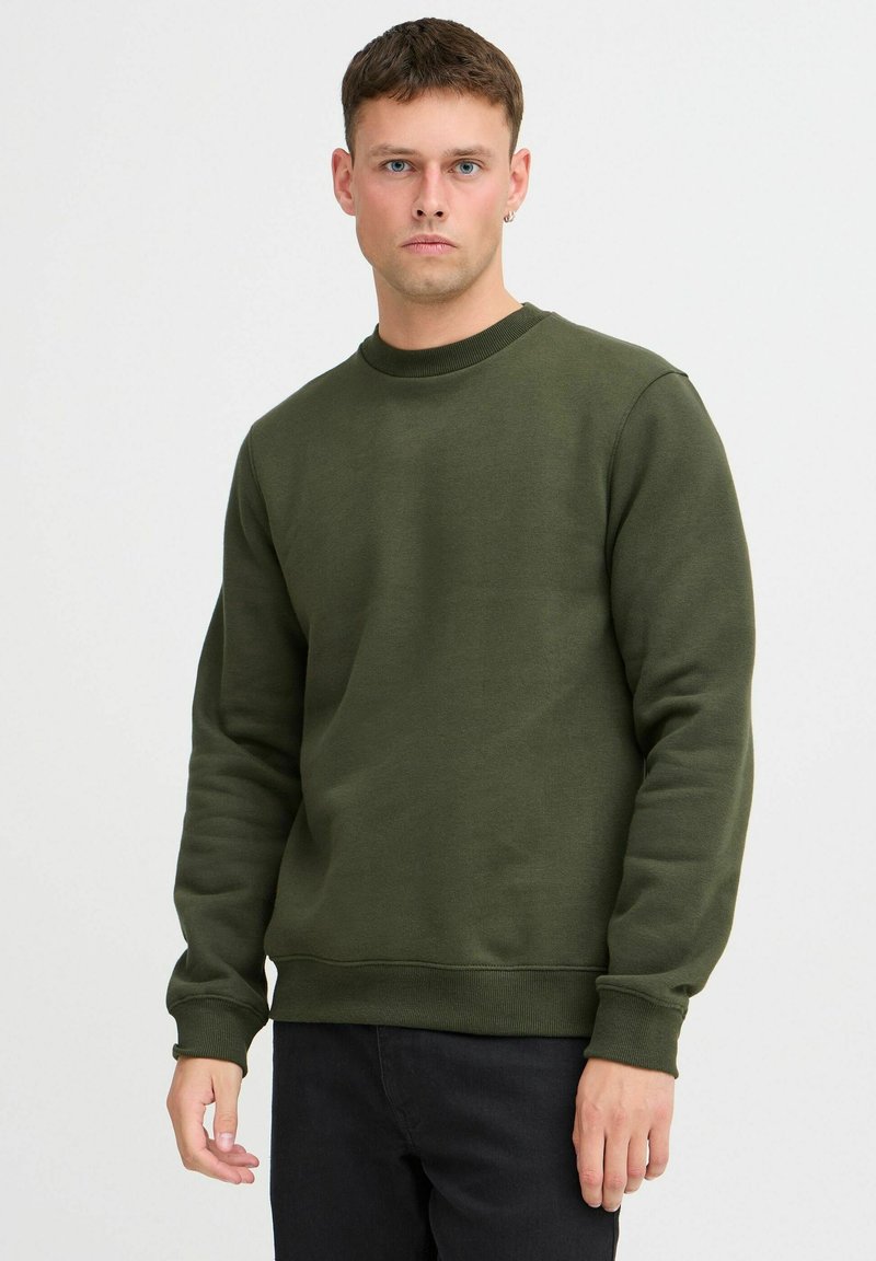 Olivengrønn genser, rund hals, lange ermer, ribbestrikkede mansjetter og kant, myk tekstur, figursydd design, brukt med mørke jeans.